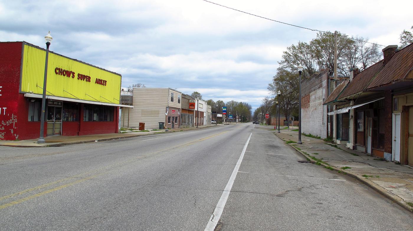 An empty four-lane main street in Rosedale, Mississippi, a painted grocery on one side and vacant shopfronts on the other